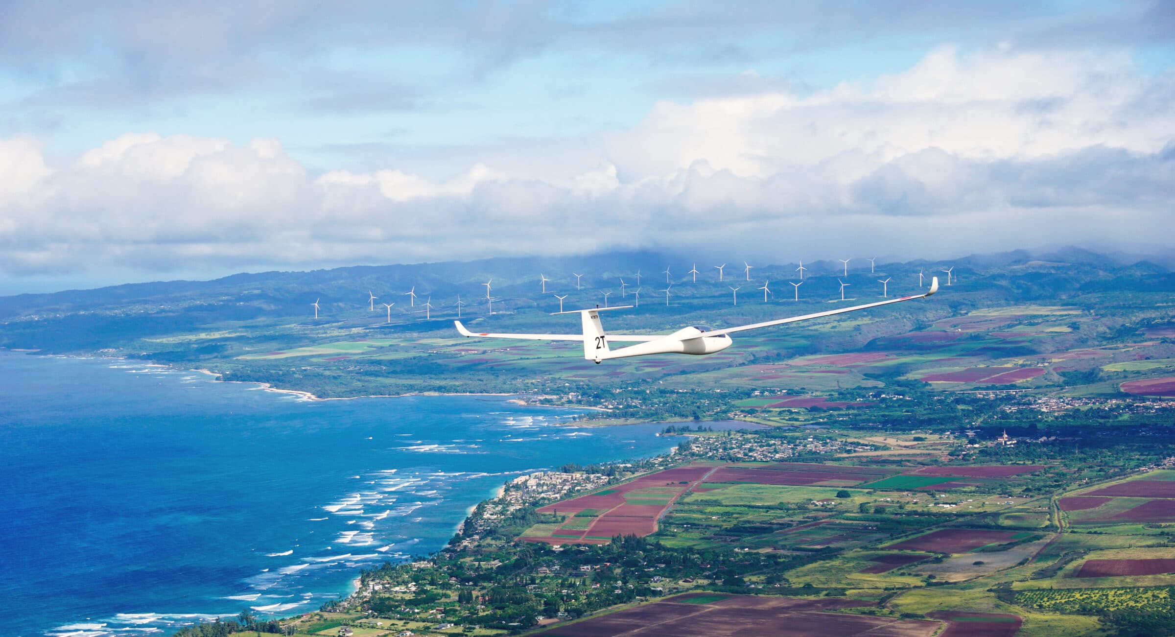 Glider gliding above the turquoise North Shore coastline past the Kahuku windmills